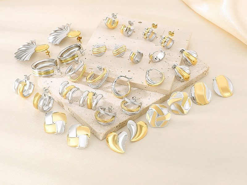 A collection of elegant gold-plated and silver-tone earrings displayed on textured stone slabs. The assortment includes various hoop, stud, and geometric designs, combining gold and silver finishes for a stylish and sophisticated look.