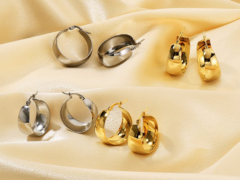 A collection of gold and silver stainless steel hoop earrings displayed on luxurious champagne-colored satin fabric. The earrings have a polished and reflective finish, showcasing their elegant and timeless design.