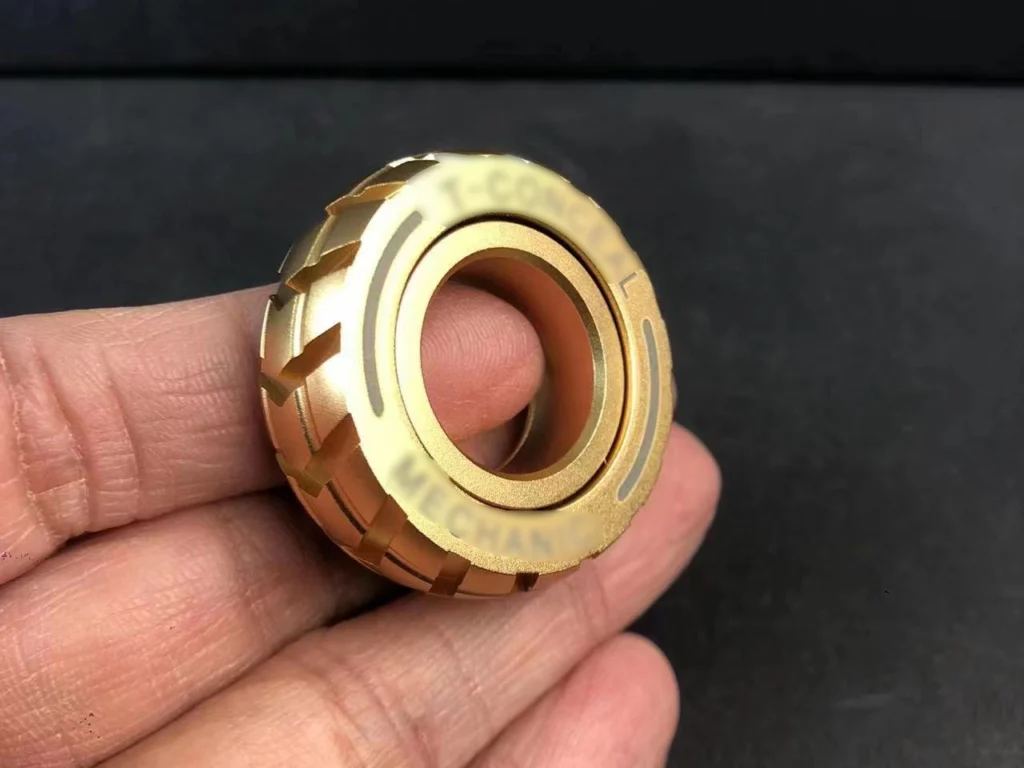 A close-up of a gold-colored fidget ring held between fingers, featuring a mechanical and textured design for stress relief and focus enhancement.