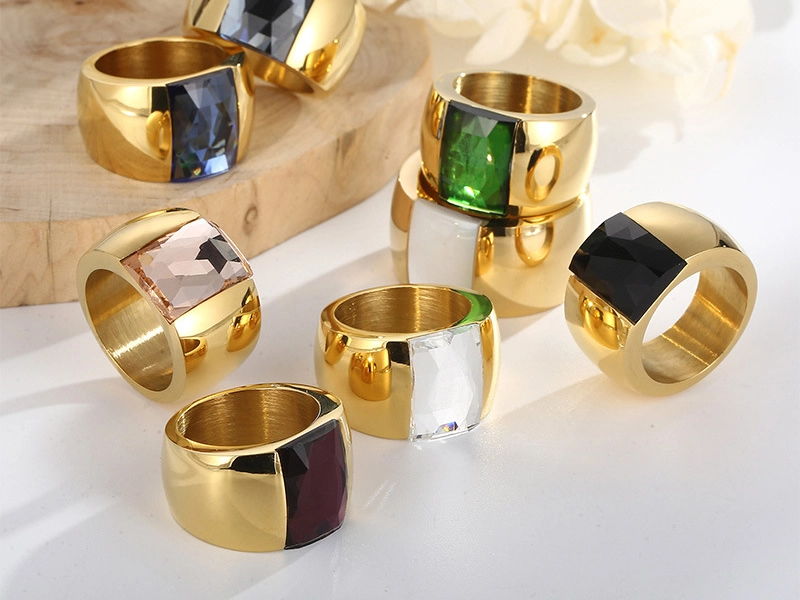 A collection of bold stainless steel rings with PVD gold coating, featuring faceted gemstone inlays in various colors.