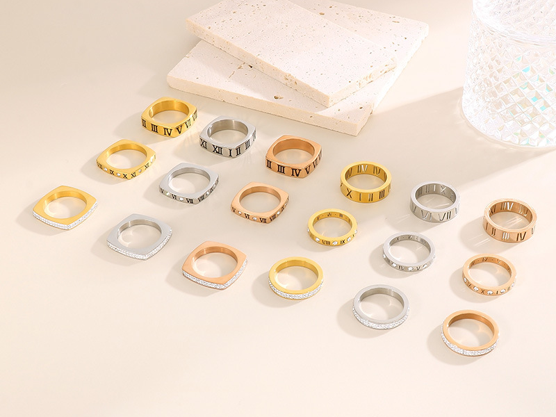 Assorted stainless steel rings with Roman numerals and crystal details in gold, silver, and rose gold finishes, arranged neatly on a beige background