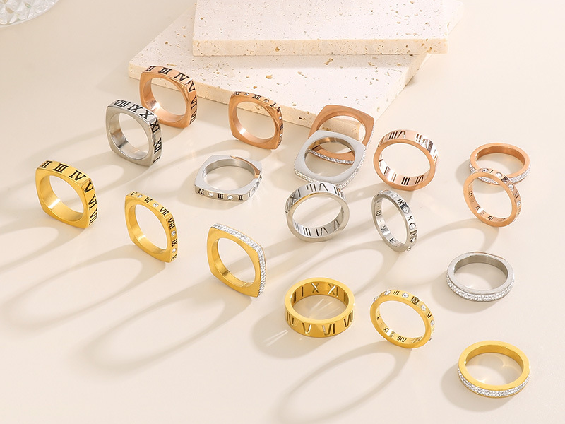 Assorted stainless steel rings in gold, silver, and rose gold finishes featuring Roman numeral engravings and crystal accents, arranged on a beige background