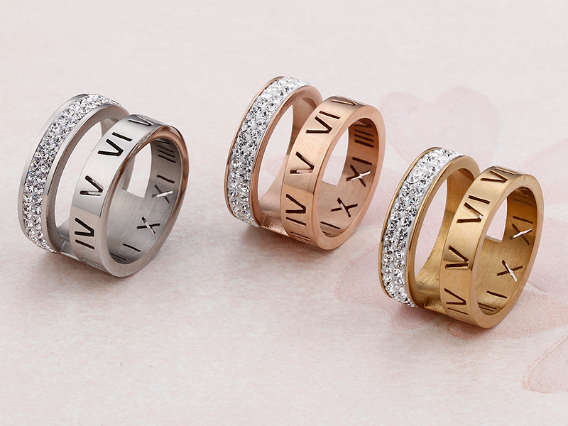 Trio of wide Roman numeral rings with pavé crystal bands in silver, rose gold, and gold finishes, displayed on a pink textured surface