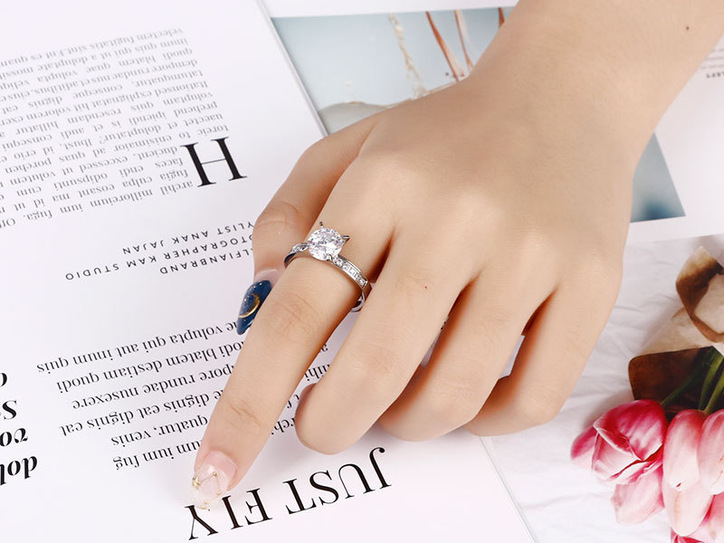 A silver solitaire engagement ring with side stones worn on a woman's hand, placed over a magazine with floral decor.